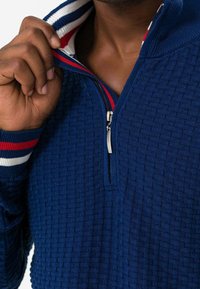 Pullover blu navy con un motivo testurizzato, zip a doppio verso e colletto a coste con dettagli a righe rosse e bianche.