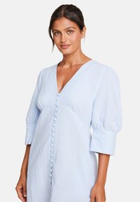 Lys blå lin kjole med V-hals, puffede ærmer og knapdetaljer foran. Glat tekstur med skrå sømme og en afslappet pasform.
