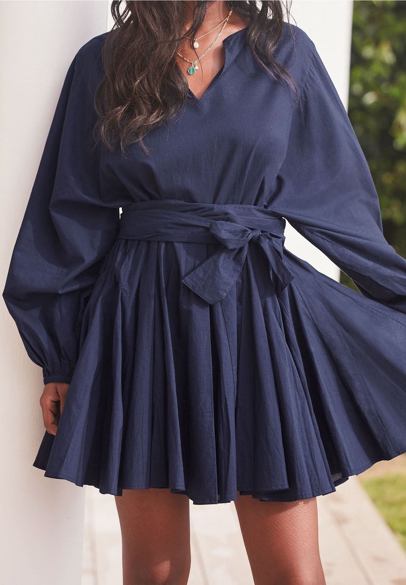 Femme portant une robe plissée bleu marine avec de longues manches bouffantes et une ceinture nouée à la taille, montrant des colliers en or avec des pendentifs.