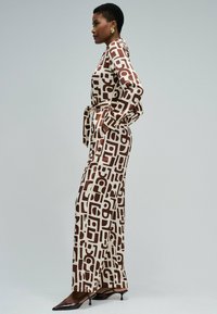 Donna in tuta a maniche lunghe con motivo a forme geometriche marroni, mani nelle tasche, indossa orecchini d'oro e tacchi neri appuntiti.