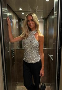 Top argentato con paillettes e collo alto, dalla forma aderente, abbinato a pantaloni neri. Sullo sfondo si possono vedere pannelli metallici di un ascensore e un'illuminazione soffusa.