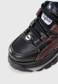 Sneaker in pelle nera con accenti rossi, suola testurizzata, piattaforma spessa e dettaglio del logo rotondo sul lato; presenta lacci robusti e cuciture.