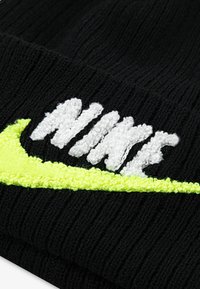 Černá pletená čepice s vyvýšeným bílým logem "NIKE" a neonově žlutým akcentem v podobě swooshu vpředu. Měkká textura a žebrovaný design.