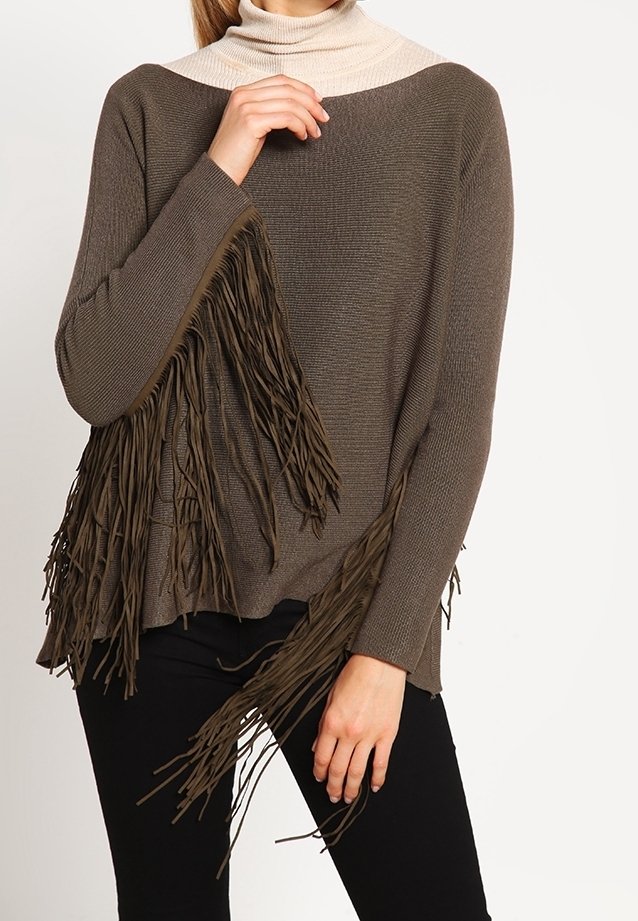 Femme portant un pull en maille marron à franges par-dessus un col roulé beige, associé à un pantalon noir, posant avec une main près du visage.