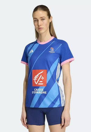 Žena v modrém sportovním dresu Francie s růžovým lemováním a logem Caisse d'Epargne, v kombinaci s tmavě modrými sportovními šortkami, stojí vzpřímeně.