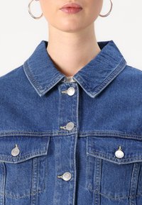 Jaqueta de denim com colarinho clássico, fecho frontal com botões, dois bolsos no peito, costuras visíveis e botões em tom prateado, na cor azul médio.