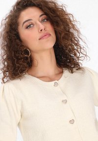 Krém színű kötött kardigán kerek nyakkivágással és rövid puff ujjakkal. Az elején szív alakú, strasszköves gombokkal díszítve.