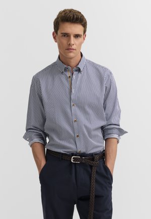 Mann trägt blau-weiß vertikal gestreiftes Hemd mit hochgekrempelten Ärmeln, marineblaue Hose und geflochtenen braunen Gürtel, Hände in den Taschen.