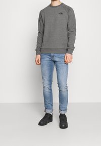 Uomo che indossa un felpa a maniche lunghe grigia della North Face, jeans slim azzurri arrotolati, calze bianche della Nike e sneakers nere su sfondo bianco.