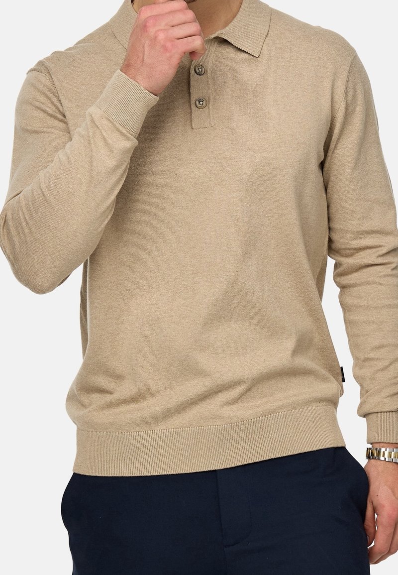 Homme portant un polo beige à manches longues avec patte de boutonnage et un pantalon bleu marine, ajustant le col avec la main gauche, montre en or au poignet droit.