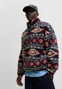 Sudadera de forro polar en azul marino, rojo y crema con patrones geométricos, cuello alto y media cremallera. Superpuesta sobre una camisa de color azul claro.