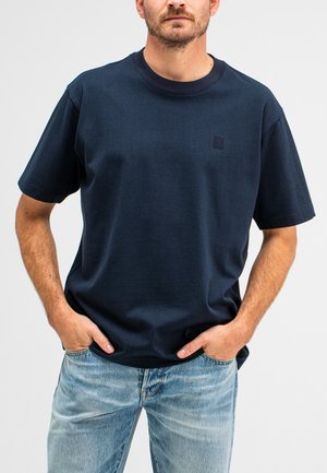 Navy blue short-sleeve t-shirt made of cotton. Features a round neck and a small logo on the left chest area. Worn with faded blue jeans.