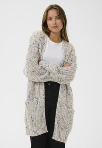 Frau mit langen braunen Haaren, die ein weißes Hemd, eine bunt gestrickte Strickjacke und schwarze Hosen trägt, steht mit verschränkten Armen vor einem weißen Hintergrund.