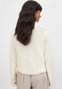 Pull en tricot crème avec un motif texturé en rayures, comportant des manches longues et une taille côtelée, porté sur un pantalon beige clair.