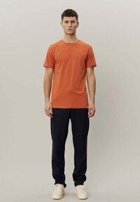 Oransje bomulls t-skjorte med rund hals, korte ermer og en subtil logo på brystet, kombinert med mørke bukser og hvite joggesko.