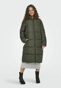 Lange olijfgroene pufferjas met een rits aan de voorkant, grote gewatteerde secties en zijkanten, gedragen met een grijze rok en zwarte haklaarzen.