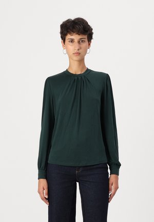 Langermet, mørkegrønn bluse laget av mykt stoff, med rund halslinning og plisserte detaljer på forsiden, kombinert med mørke denimjeans.
