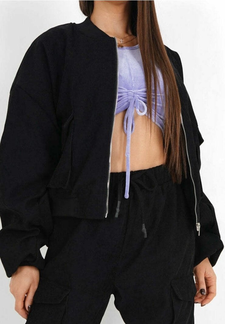 Vrouw in een zwarte ritssluiting jas over een gerimpeld lavendel crop top en zwarte cargobroek, met lang steil bruin haar.