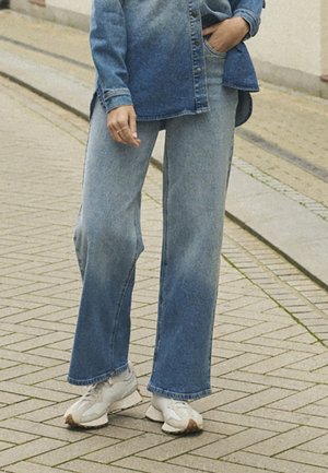 Persona vestida con jeans de pierna ancha descoloridos y zapatillas blancas, de pie sobre una acera de baldosas, con la mano apoyada en el bolsillo.