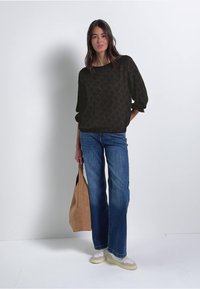 Zwarte blouse met patroon, blauwe wijde jeans, lichte schoenen en een tan tote bag. Model staat tegen een witte achtergrond.