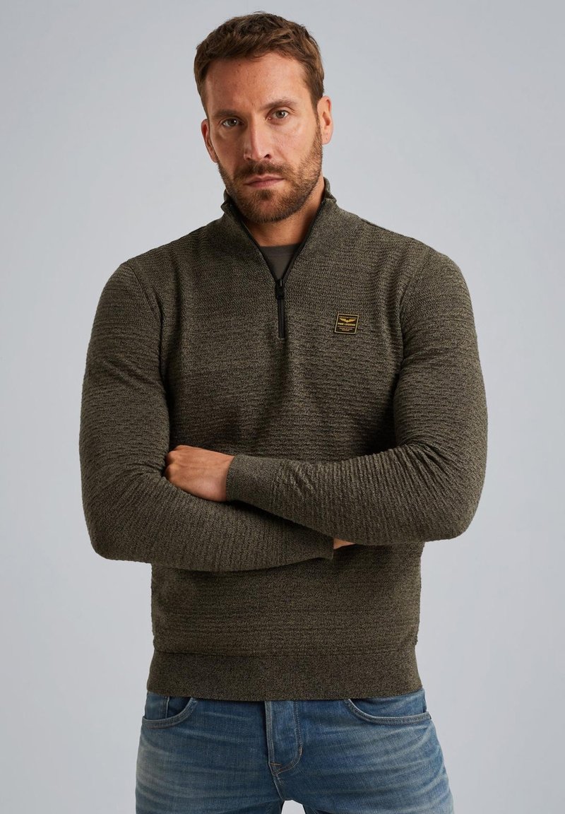 Olijfgroene zip-up trui met een gestructureerd patroon, lange mouwen en een klein logo-patch op de borst. Draag met blauwe jeans.