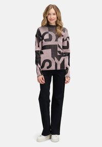Rosa Strickpullover mit großen schwarzen Buchstaben, ausgestattet mit einem gerippten Textur und einem Rundhalsausschnitt. Kombiniert mit schwarzen weit geschnittenen Hosen und weißen Schuhen.