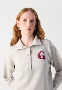 Grå tröja med krage, försedda med guldfärgade tryckknappar och en bordeauxbroderad "G". Mjuk tyg med en avslappnad passform.