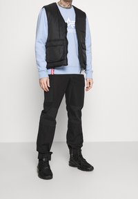 Gilet imbottito nero sopra una felpa azzurra chiara, abbinato a pantaloni cargo neri con molteplici tasche e cerniere, completati da sneaker nere.