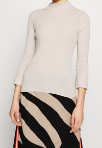 Donna che indossa un maglione a dolcevita beige a coste e una gonna nera con strisce ondulate beige e strisce rosse laterali, in piedi davanti a uno sfondo bianco.