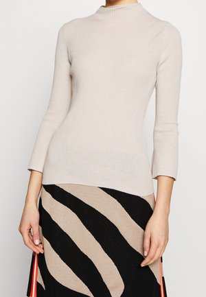 Kvinde iført en beige ribbet højhalset sweater og en sort nederdel med beige bølgestreger og røde sidelister, der står foran en hvid baggrund.