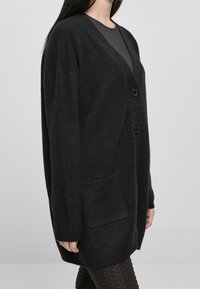 Cardigan en tricot noir avec un col en V, deux boutons, une poche avant et des poignets côtelés. Texture douce et coupe légèrement oversize.