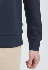 Marine sweatshirt med ribbet manchet og kant, lavet af et tekstureret stof. Grøn etiket synlig på siden, parret med beige fløjl bukser.