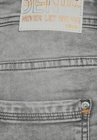 Tessuto denim grigio con un'etichetta cucita che riporta la scritta "DENIM NEVER LET ME GO CECIL", con accenti arancioni e un dettaglio di bottone metallico.