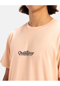 Camiseta de color melocotón hecha de tejido suave, con un logo impreso en negro en el pecho que dice "Quiksilver The Original Surf Co Since 1969".