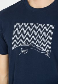 Marinblå bomullströja med en vit fiskdesign och vågiga linjer ovanför, som skapar en marin-tema grafik. Rund halsdesign.