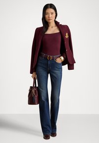 Γυναίκα όρθια, φοροντας μπορντό σακάκι πάνω από ταιριαστό τοπ, μπλε τζιν, μπορντό ζώνη και τσάντα, χρυσό βραχιόλι και κρίκους σκουλαρίκια σε λευκό φόντο.