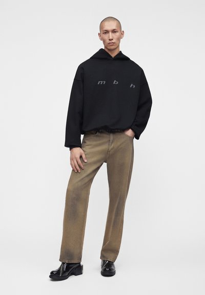 Homme portant un sweat à capuche noir avec le texte "mbh", un jean marron délavé droit et des bottines noires à talons, debout, une main dans la poche.