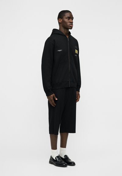 Μαύρο φούτερ με φερμουάρ μπροστά και τσέπες, με λογότυπο, συνδυασμένο με μαύρο σορτς μέχρι το γόνατο και μαύρα παπούτσια με λευκές κάλτσες.