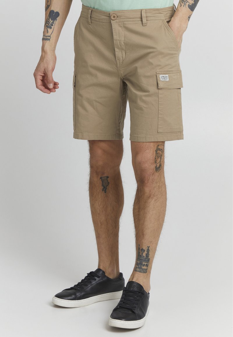Khaki spodenki cargo o prostym kroju, z bocznymi kieszeniami i teksturowanym materiałem. Połączone z czarnymi sneakersami w niskiej wersji.