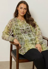 Fließende Bluse mit langen Ärmeln im grünen und lila Batikmuster, ausgestattet mit einem vorderen Bindedetail, Falten und einer leichten Textur.