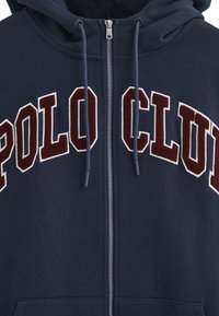 Felpa con zip colore navy in tessuto felpato, con la scritta "POLO CLUB" in bordeaux e bianco. Include cordini e dettaglio della zip in metallo.