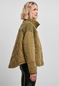 Olivenfarget quilted jakke med hette, med en myk, teksturert krage og en boksaktig silhuett. Stoffet er glatt med en matt finish.