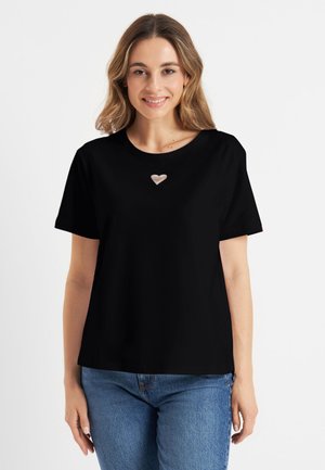 Młoda kobieta uśmiechająca się, ubrana w czarny T-shirt z małym wycięciem w kształcie serca na piersiach i niebieskie jeansy, stojąca na białym tle.