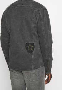 Hiilenharmaa takki, jossa kulunut pinta, musta merkki vihreällä tähdellä, näkyvät kontrastitikkaukset ja vetoketjulla varustetut hihansuut.