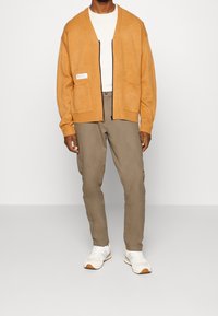 Homme portant un cardigan zippé moutarde sur une chemise blanche, un pantalon vert olive et des baskets blanches avec des accents bleu clair.