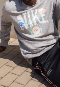 Person som bär en grå Nike-tröja med färgglad logotyp och svarta Nike-byxor, sitter på asfalterad mark utomhus.