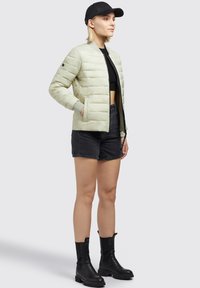 Light green padded jacket with a bomber design, ribbed cuffs, front pockets, and black accents, worn with black shorts and knee-high boots.