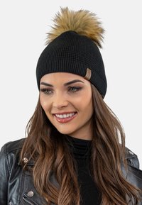 Zwarte gebreide beanie met een gestructureerd patroon, voorzien van een verwijderbare tan nepbont pompon bovenop. Inclusief een klein leren label aan de zijkant.