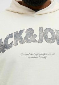 Kremowa bluza z kapturem z dużym, teksturowanym logo "JACK & JONES" w ciemnoszarym kolorze oraz mniejszym hasłem poniżej, wykonana z miękkiego materiału bawełnianego.