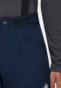 Granatowe spodnie narciarskie mają boczną kieszeń z zamkiem błyskawicznym, regulowany pas na rzepy oraz logo na nogawce. Wykonane z trwałej, teksturowanej tkaniny.